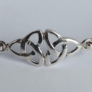 May include: A polished silver bracelet featuring a central focal point composed of two mirrored triquetra knots. The metal is reflective and smooth, with the knot design connected to thin, curved silver bands on either side via small circular links.