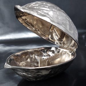 May include: A silver-toned metal nutcracker shaped like a walnut shell. The object is hinged and opens to reveal a textured interior with two serrated metal teeth designed for cracking nuts. The exterior surface mimics the rough texture of a walnut.
