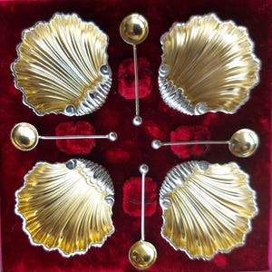 Pode incluir: Um conjunto de quatro saleiros de prata em forma de concha com colheres correspondentes. As conchas são banhadas a ouro e têm um design nervurado detalhado. As colheres têm cabos redondos em forma de bola.