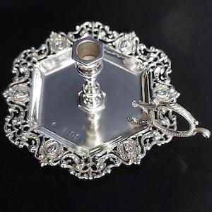 May include: A silver-colored metal chamberstick candle holder featuring a hexagonal base with an intricate, openwork filigree border. The central candle socket is hexagonal, and a small, decorative handle is attached to one side of the base.