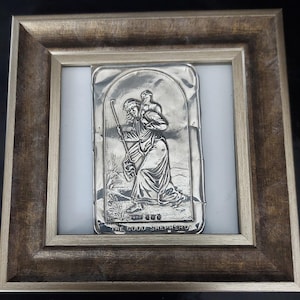 May include: A rectangular silver-toned metal plaque featuring a relief of a figure carrying a sheep, titled THE GOOD SHEPHERD at the bottom, set within a square, distressed metallic gold-toned frame with a white border surrounding the plaque.
