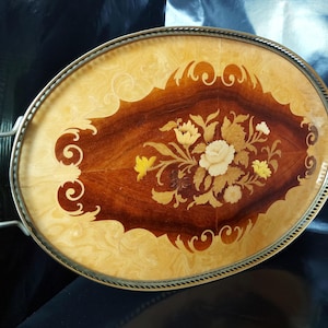 May include: An oval serving tray featuring a central wood inlay design of flowers in shades of cream, yellow, and brown. The tray has a light-coloured wood border, a dark wood centre, and a metallic rim with two side handles for easy carrying.