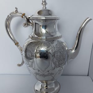 Puede incluir: Una tetera de plata con un diseño ornamentado y una tapa. La tetera tiene un cuerpo redondo y un pico curvo. El asa es curva y tiene un diseño decorativo.
