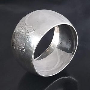 Puede incluir: Un servilletero de plata pulida con un monograma floral grabado. El anillo es circular con una superficie lisa y reflectante. El monograma está en la superficie exterior del anillo. El fondo es oscuro.