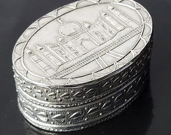 May include: Silver oval trinket box with an embossed design of the Taj Mahal on the lid. The box has a decorative border around the edge.