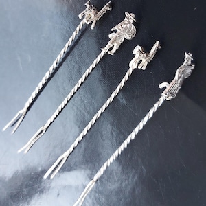 May include: Four silver cocktail stirrers with twisted stems and decorative tops. The tops feature figures of a llama, a person in a hat, and a person with a shield. The stirrers have a two-pronged end.