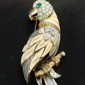 Peut inclure: Broche vintage en forme de perroquet, en métal doré, avec émail crème, gris et blanc. La tête du perroquet est ornée de strass transparents et d'un œil vert. L'oiseau est perché sur une branche, avec une fermeture à épingle au dos.