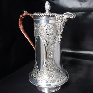 May include: A silver pitcher with intricate floral and scrollwork engravings. The pitcher has a curved spout, a lid with a decorative finial, and a handle wrapped in a brown material. The design is ornate and detailed.