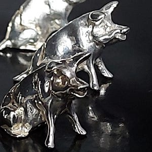 May include: Two small, reflective silver-toned pig figurines are positioned one behind the other. Each pig is depicted in a standing posture with detailed facial features, including pointed ears and a snout, captured in a polished, metallic finish.