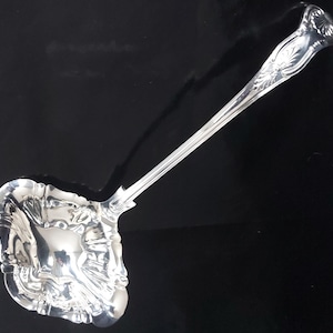 May include: A silver-colored metal ladle featuring a square-shaped bowl with scalloped, decorative edges and a long, slender handle. The handle terminates in an ornate, shell-like motif with intricate embossed detailing along its length.