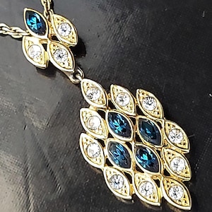以下が含まれることがあります： リーフデザインのペンダントが付いたゴールドトーンのネックレス。ペンダントには、透明と濃い青色の宝石が交互に飾られています。ネックレスは暗い背景にセットされています。