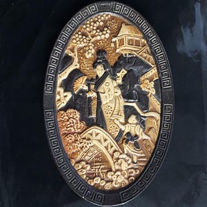 May include: An oval relief carving showing an Asian scene with figures, a bridge, and a building, set within a decorative border. The colour scheme includes shades of brown, black, and cream.