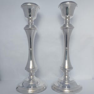 Op de afbeelding: Een paar hoge, gepolijste zilverkleurige kandelaars met een glad, taps toelopend ontwerp. Elke kandelaar heeft een brede, ronde voet met concentrische ringdetails en een uitlopende bovenkant die is ontworpen voor een enkele kaars.