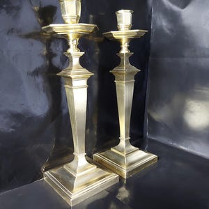 May include: A pair of antique brass candlesticks. Each candlestick features a square base, a slender, tapering stem, and a hexagonal candle cup. The candlesticks are a warm gold colour and are set against a dark backdrop.