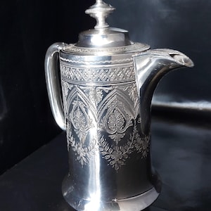Puede incluir: Una cafetera de metal plateado con un patrón grabado de motivos florales y bandas geométricas. El recipiente tiene un cuerpo cilíndrico, un asa curva, una base ensanchada y una tapa extraíble rematada con un pequeño remate decorativo.