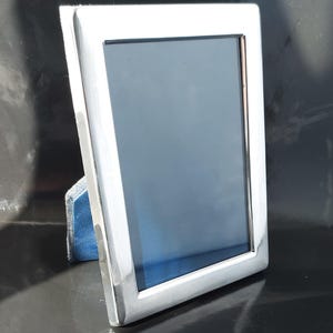 Könnte beinhalten: Ein rechteckiger, silberfarbener Bilderrahmen mit dunkelblauem Innenraum. Der Rahmen hat ein schlichtes, modernes Design mit poliertem Finish. Der Rahmen wird von einer blauen Samtunterlage gestützt.