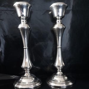 Op de afbeelding: Twee zilverkleurige kandelaars met een klassiek ontwerp. Elke kandelaar heeft een brede, ronde basis, een slanke, gebogen steel en een uitlopende bovenkant. De kandelaars zijn gemaakt van glanzend, reflecterend metaal.