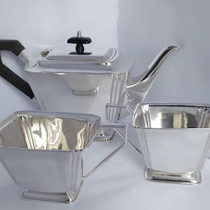 Puede incluir: Un juego de té plateado que incluye una tetera con asa y tapa negras, un azucarero y una crema. El juego tiene un diseño geométrico y se exhibe sobre una superficie blanca. La tetera mide aproximadamente 20 cm de alto.