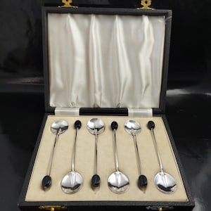 Op de afbeelding: Een set van zes zilveren lepels met zwarte ovale details, gepresenteerd in een zwarte doos met een crèmekleurig interieur. De lepels hebben een gepolijst, reflecterend oppervlak en de doos heeft goudkleurige sluitingen.