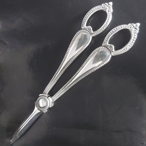 Puede incluir: Cascanueces plateado con detalles ornamentados. Los mangos tienen un diseño ovalado con un borde de cuentas, y las cuchillas son puntiagudas. El cascanueces está diseñado con una bisagra decorativa y un diseño circular en el centro.