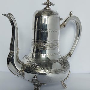 Op de afbeelding: Een zilveren koffiepot met een gebogen tuit en een decoratieve handgreep. De pot heeft een deksel met een knop en is versierd met bloemen- en geometrische patronen. Hij staat op drie sierlijke voetjes.
