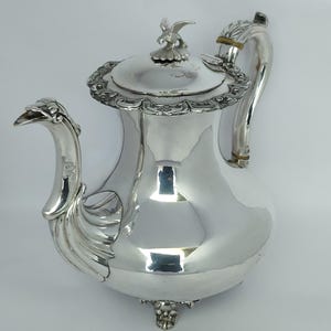 Può includere: Una teiera in argento lucido con un pomello a forma di uccello sul coperchio, un manico ornato con dettagli decorativi, un beccuccio curvo con fattezze di uccello e complessi motivi a volute lungo il bordo e la base del recipiente.
