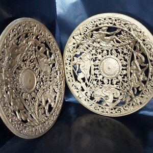 Könnte beinhalten: Zwei kreisförmige, goldfarbene Wandplatten mit komplexen, durchbrochenen Reliefdesigns. Jede Platte zeigt ein zentrales Medaillon, das von detaillierten Figuren von Putten inmitten von Ranken und floralen Mustern umgeben ist.