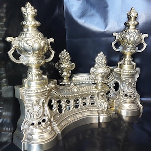 Op de afbeelding: Een paar haardbokken van gepolijst messing met hoge, sierlijke ornamenten in urnvorm en gedetailleerd krulwerk. De basis van elke haardbok heeft een gebogen, opengewerkt ontwerp met decoratieve reliëfpatronen over het gehele oppervlak.