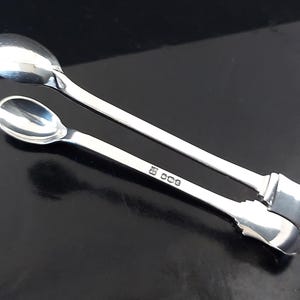 Peut inclure: Une paire de pinces à sucre en argent avec des extrémités en forme de cuillère. Les pinces sont en métal poli et ont un design simple et élégant. Les poinçons sont visibles sur l'un des bras. Les pinces sont posées sur un fond noir.