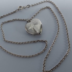 May include: A silver-toned heart-shaped locket pendant suspended from a matching silver-toned cable chain. The locket features an engraved leaf-like pattern on its front surface and a textured border, with a circular clasp visible on the chain.