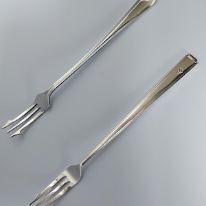 May include: Two silver-toned metal cocktail forks with long, slender handles. One fork features a decorative shell-like motif at the top of the handle, while the other displays a small, square-shaped emblem near the end of its smooth, tapered handle.