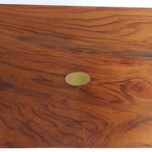 Puede incluir: Una caja de madera rectangular con un acabado marrón rojizo intenso y patrones de vetas de madera prominentes. Una pequeña placa ovalada de latón está centrada en la tapa. La caja parece hecha a mano y es adecuada para el almacenamiento.