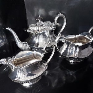 May include: A silver tea set consisting of a teapot, a creamer, and a sugar bowl. Each piece features a polished metallic finish, ornate embossed floral detailing along the sides, and scalloped edges. The teapot has a curved spout.