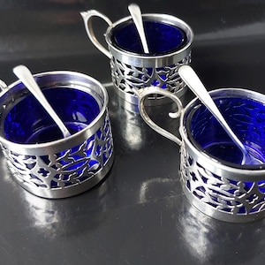 Op de afbeelding: Drie antieke verzilverde zoutvaatjes met kobaltblauwe glazen inzetstukken. Elk zoutvaatje heeft een delicaat, opengewerkt ontwerp en een klein lepeltje. De zoutvaatjes hebben handvatten en zijn op een donkere ondergrond geplaatst.