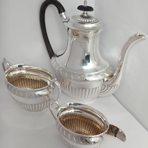 Pode incluir: Um conjunto de chá em prata composto por um bule com pega escura curva e um açucareiro e leiteira a condizer. Cada peça apresenta um acabamento em prata polida, uma base canelada e uma faixa decorativa com um padrão geométrico.