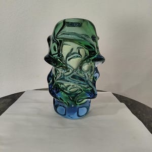 May include: A hand-blown glass vase featuring an irregular, organic shape with a swirling color gradient that transitions from a deep forest green at the top to a vibrant sky blue at the base, characterized by thick, translucent glass walls.