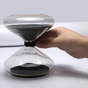 May include: A glass hourglass with black sand. The hourglass is being held by a hand.