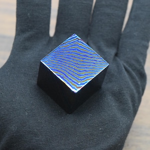 Puede incluir: Un pequeño objeto negro cuadrado con una parte superior estampada. La superficie superior presenta un diseño ondulado azul y dorado. El objeto se sostiene con un guante negro.