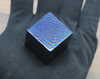 May include: A small, square, black object with a patterned top. The top surface features a blue and gold wave-like design. The object is held in a black glove.