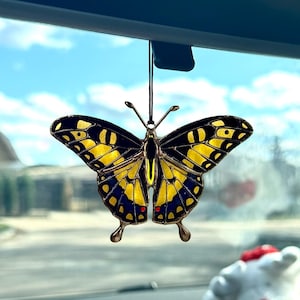 May include: A decorative butterfly car charm with yellow and blue wings hangs from a rearview mirror. The butterfly has a detailed design and is suspended by a thin string. Sunglasses and a small plush toy are on the dashboard.