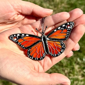 May include: A stained glass butterfly with orange and black wings, and white dots along the edges. The butterfly is held in open hands against a blurred green background. The butterfly's body is black.