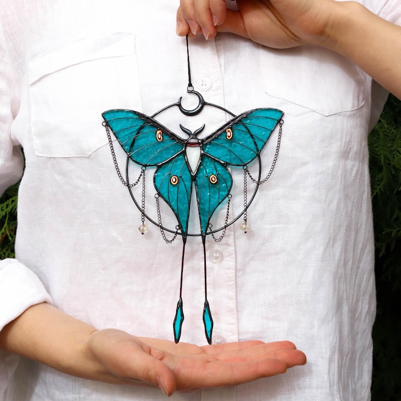 May include: A teal blue stained glass luna moth hanging from a black metal chain with a crescent moon design. The moth has a white body and black accents. The chain has small clear beads hanging from it.