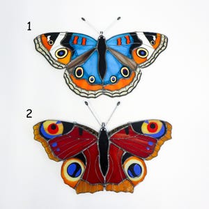 May include: Two stained glass butterfly decorations. The top butterfly has blue, orange, and black wings. The bottom butterfly has red, yellow, and black wings. Both have detailed patterns and are set against a white background.