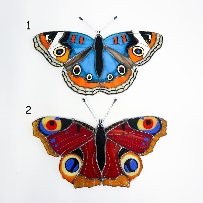 May include: Two stained glass butterfly decorations. The top butterfly has blue, orange, and black wings. The bottom butterfly has red, yellow, and black wings. Both have detailed patterns and are set against a white background.