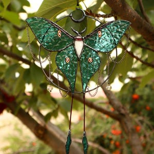 May include: A stained glass moon moth hanging ornament with a crescent moon and chains. The moth is green with iridescent wings and a white body.