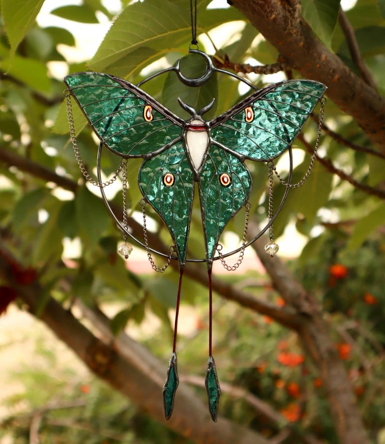 May include: A stained glass moon moth hanging ornament with a crescent moon and chains. The moth is green with iridescent wings and a white body.