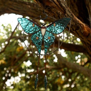 May include: A teal blue stained glass luna moth hanging from a branch. The moth has a crescent moon design and dangling chains with crystal beads.