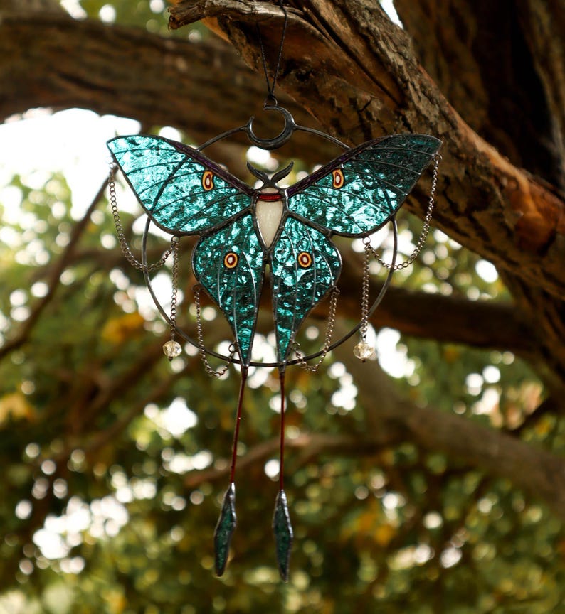May include: A teal blue stained glass luna moth hanging from a branch. The moth has a crescent moon design and dangling chains with crystal beads.