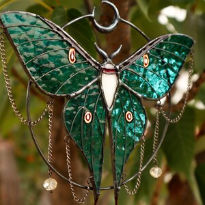 May include: A stained glass moon moth with green wings, red and white accents, and a crescent moon design. The moth is suspended from a metal ring with chains and crystal beads.