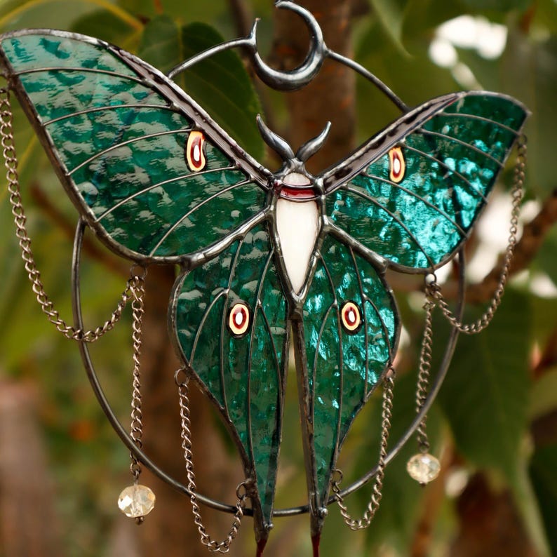 May include: A stained glass moon moth with green wings, red and white accents, and a crescent moon design. The moth is suspended from a metal ring with chains and crystal beads.